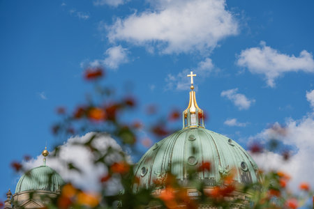 A cathedral dome topped with a golden cross framed by blurred orange flowers. The blue sky with clouds emphasizes the architecture s grandeur.の写真素材