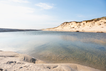 A tranquil stream of seawater cuts through the sandy shore beneath grassy dunes. The bright sunlight enhances the colors of sea and sand.の写真素材
