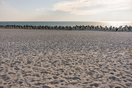 Sunlight reflects on the calm sea behind a wide sandy beach lined with concrete wave breakers. The scene feels peaceful and minimalistic.の写真素材