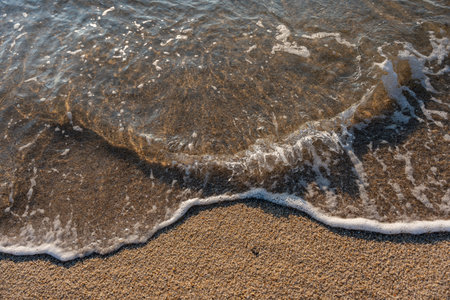 Clear water ripples over wet sand as a wave retreats, sparkling in the sun. The texture of the beach...