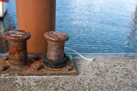 Two rusted bollards on a concrete pier with ropes attached. The textured metal contrasts with the blue water in the background.の写真素材