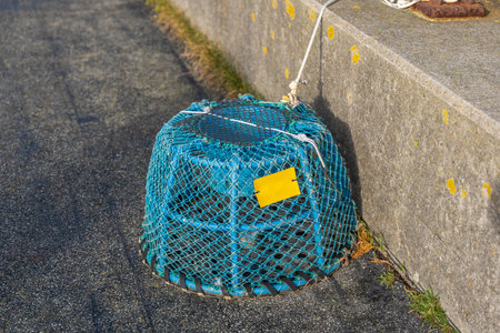 A blue fishing trap covered with a net and tied with rope sits beside a concrete pier wall. The bright yellow tag adds contrast to the marine equipment.の写真素材