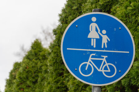 Blue pedestrian and bicycle sign indicates a shared path for walkers and cyclists. The background shows green hedges under a cloudy sky.の写真素材