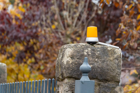 Orange warning light mounted on a stone fence pillar. Autumn leaves blur in the colorful background.