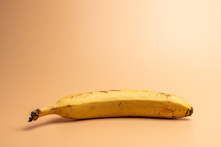 A ripe banana lies horizontally on a warm beige surface. The banana shows natural spots and a gentle curve.の写真素材