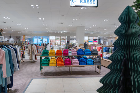 Germany Berlin December 3, 2025. Colorful folded sweaters on display beneath a checkout sign. Minimalist department store interior with holiday decor and a stylized tree sculpture.のeditorial素材