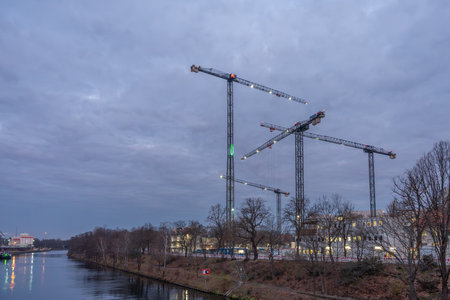 Construction cranes rise over a riverside worksite beneath a moody twilight sky. The canal and bare...