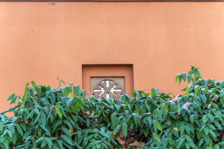 A decorative stone ventilation block is framed by green foliage against a warm-colored wall. The image combines architectural detail with natural elements.の写真素材