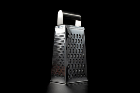 Stainless steel box grater standing on a black background with dramatic studio lighting. Clean,...