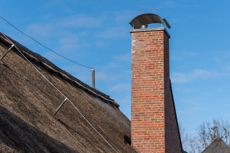 A brick chimney with a metal cap on top. The chimney is on a roof. The roof is made of strawの写真素材