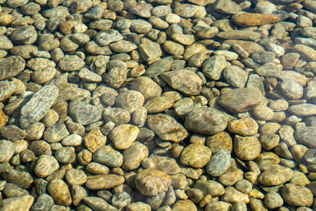 Clear water flows over smooth, rounded river stones. Sunlight glints off the pebbles in shallow...