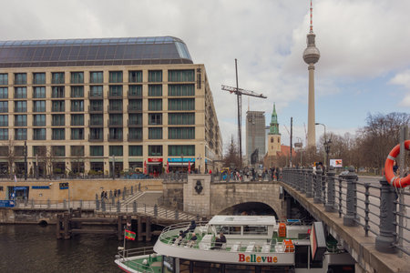 Germany Berlin March 27, 2026. Bridge spans river, connecting urban buildings. Clouds hover above Berlin's skyline. Tourist boat glides beneath stone arches. Tall tower rises behind residential structuresのeditorial素材