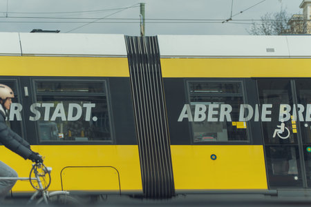 Germany Berlin March 27, 2026. Yellow tram with bold white lettering passes by. Cyclist waits beside the curb, helmeted and calm. Urban scene under overcast sky, quiet and routine. Tram's side reads "Stadt". Perfect for city lifeのeditorial素材