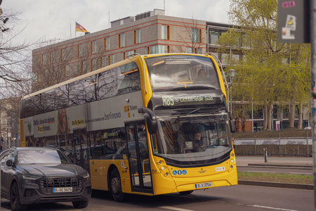 Germany Berlin March 27, 2026. Yellow double-decker bus moves through urban street. Car passes...