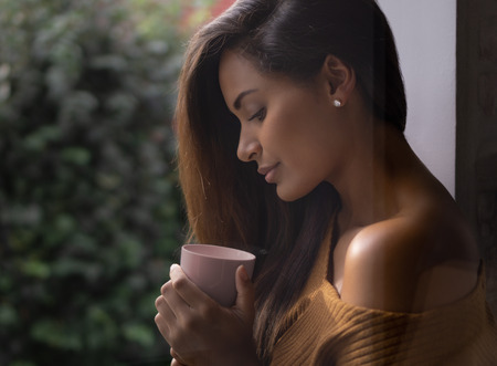 Young beautiful model holding a warm cup of coffee in the morning at a window.の写真素材