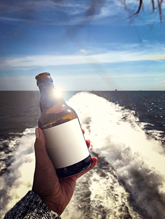 A blank labeled brown glass bottle held in a female hand in front of a water splash made by a jet stream of a ferry boat.の写真素材