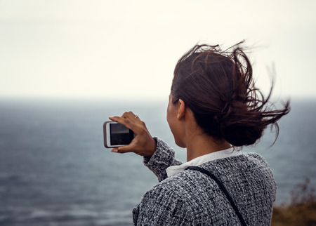 Beautiful young woman taking photos and selfies with her smartphone of the ocean with wind going through her long hair.の写真素材