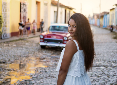Beautiful dark skinned young lady in white dress standing in front of an old classic car in the old streets of Trinidad in Cuba.の写真素材