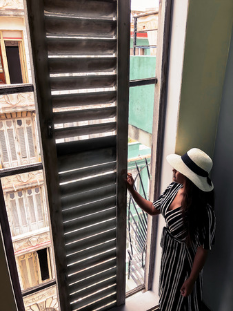 Young female model posing in beautiful black and white dress and hat looking out an old wooden window in Havana Cuba. Old balconies and windows are to be seen in the background.の写真素材