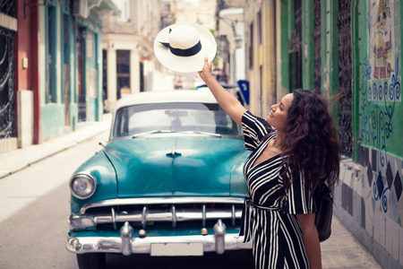 Young beautiful black woman with a typical cuban hat standing in the old streets of Havana Cuba in front of a classic car holding the hat right over the car.の写真素材