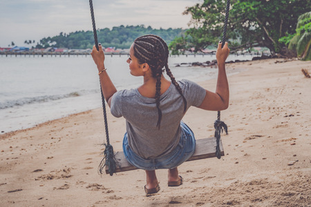 Young female from behind with braided hair sitting on a swing at a beach in Thailand.