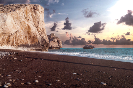 View at the AproditeÂ´s rock in Paphos, Cyprus with a cloudy sunset and the ocean with waves in the background. Dark sand with pebbles and rocks is to be seen in the foreground.の写真素材