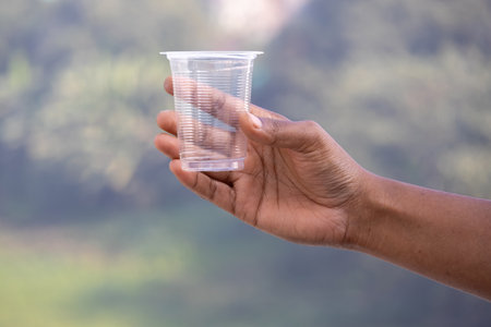 A female hand holds an empty plastic cup against a blurred nature background, illustrating the...