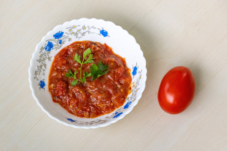 Delicious Tomato Bhorta is served in a white ceramic bowl on a light wooden surface, with a whole red tomato resting beside it. Tomato bharta is a popular Bengali dish.の写真素材