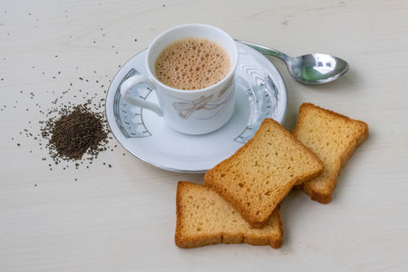 Hot, frothy milk tea in a teacup with golden rusk biscuits. Fresh and dried tea leaves scattered on a wooden surface, capturing the essence of a cozy beverage experience.の写真素材