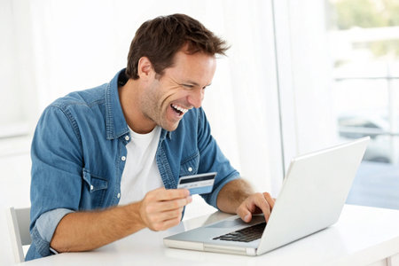 A man in a denim shirt laughs while holding a credit card and typing on a laptop computer.の素材