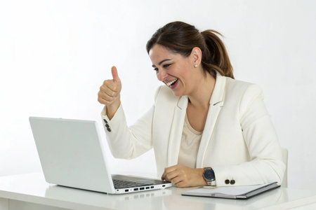 A happy woman in a white blazer gives a thumbs up while looking at her laptop, indicating achievement.の素材