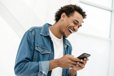 A happy young man with curly hair laughs heartily while holding and looking at his mobile phone.の素材
