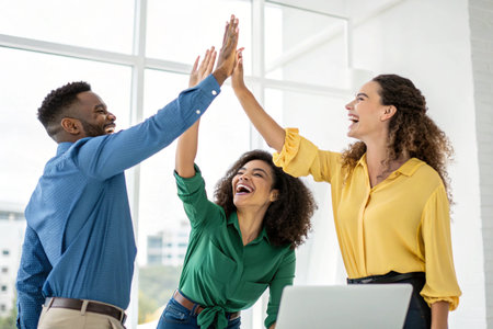 Three diverse colleagues enthusiastically high-fiving, smiling, and laughing, symbolizing teamwork and achievement in a modern workspace.の素材
