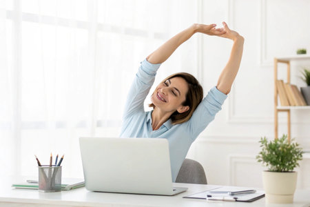 A woman in an office setting stretches her arms above her head, taking a break from her work.の素材