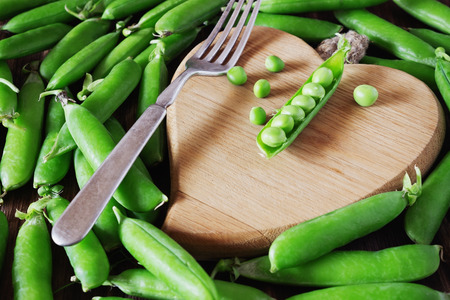 Pods of fresh green peas and wooden cutting board in the shape of a heart. Healthy food concept. Organic vegetables. Selective focusの写真素材