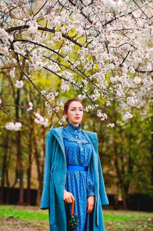 portrait of a redhaired young woman in a blue coat and retro dress in a rainy spring garden of apple trees. Spring time.の写真素材