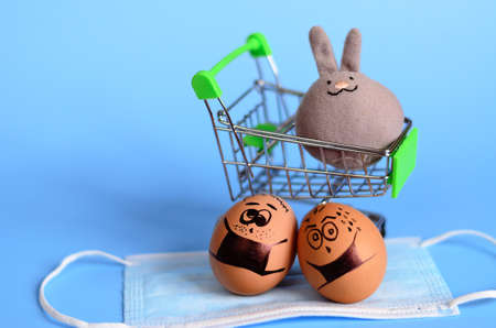 Creative Easter eggs with Corona virus (COVID19) protection concepts. Diverse chicken eggs on mini shopping cart with doodle faces wearing medical masks on blue background. Flat lay, top view.の写真素材