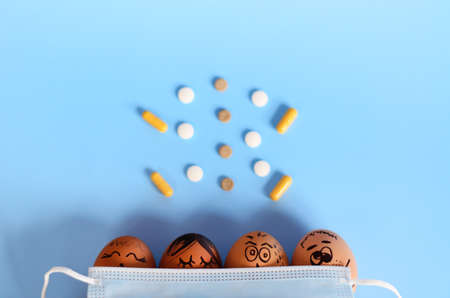 Creative Easter chicken eggs with doodle faces wearing medical masks with pills on blue background. Flat lay, top view, mockup, copy space.の写真素材