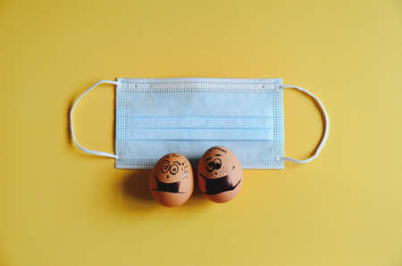 Creative Easter chicken eggs with doodle faces wearing medical masks on yellow background. Flat lay, top view, mockup, copy space.の写真素材