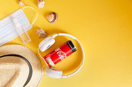 PortoroÅ¾. 02.02.2021.Travel under Covid-19 and new normal concepts. Top view of white headphones, can of Coca- Cola, phone and beach hat on yellow background. Creative ideas of prevent coronavirus.のeditorial素材
