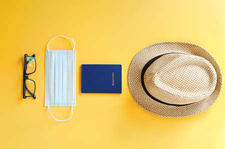 Top view of medical face mask, hand sanitizer, passport, headphones and beach hat on yellow...