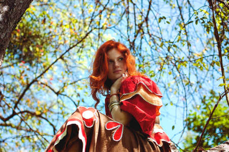 Stylish girl with freckles and red hair in red long dress holding red poppy flower at green field on nature. Portrait of boho woman posing in summer countryside. Happy lifestyle. Atmospheric moment.の写真素材