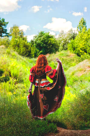 Stylish girl with freckles and red hair in red long dress dancing at green wild forest on nature. Portrait of boho woman posing in summer countryside. Happy lifestyle. Atmospheric momentの写真素材