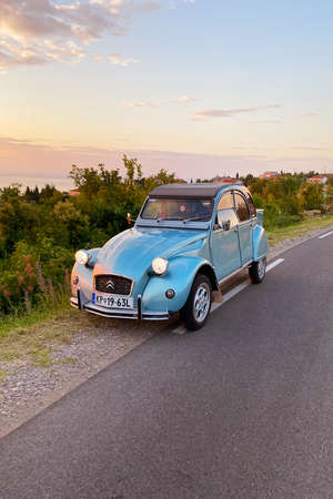 Beautiful blue retro car Citroen stands on the road against red sunset at the Adriatic sea, coast in Slovenia at summer. View to green hills and olive trees.のeditorial素材