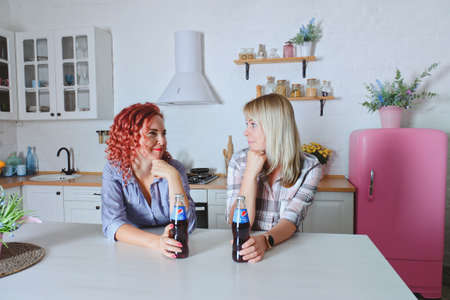 Young cute women, sisters, friends 30+ in blue jeans and a shirt sitting and relaxing in bright clean kitchen. Drinking soda. Bottles of Pepsi at the table.のeditorial素材