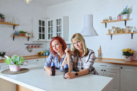 Young cute women, sisters, friends 30+ in blue jeans and a shirt sitting and relaxing in bright clean kitchen. Drinking soda. Bottles of Pepsi at the table.のeditorial素材