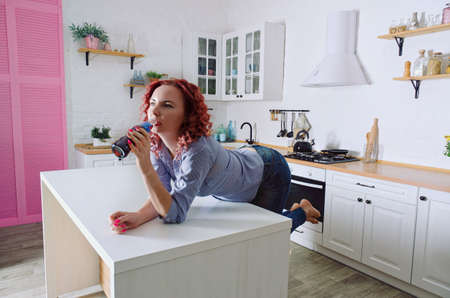 Young cute redhead woman 30+ in blue jeans and a shirt sitting and relaxing in her bright clean kitchen. Drinking soda. Bottle of pepsi at the tableのeditorial素材