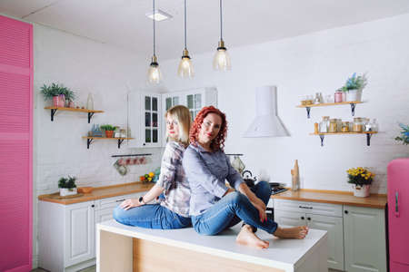 Young cute women, sisters, friends 30+ in blue jeans and a shirt sitting and relaxing in bright clean kitchen.の写真素材