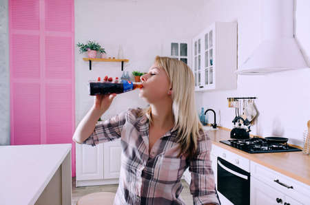 Young cute woman 30+ in blue jeans and a shirt sitting and relaxing in her bright clean kitchen. Phone in hand. Bottle of pepsi at the tableのeditorial素材