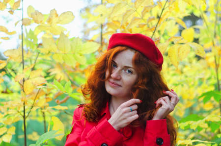Young happy woman with red hair, freckles, blue eyes in a red beret, coat walking in autumn park, smiling. Yellow fall leaves.の写真素材